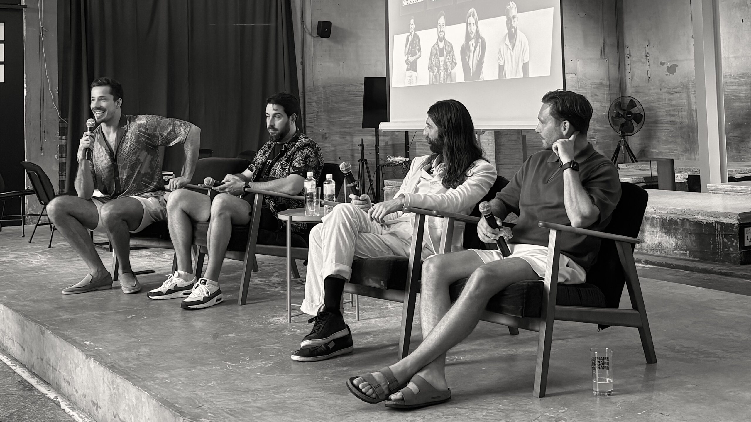 Four casually dressed men engage in a relaxed panel discussion in a modern industrial-style venue with a presentation screen in the background.