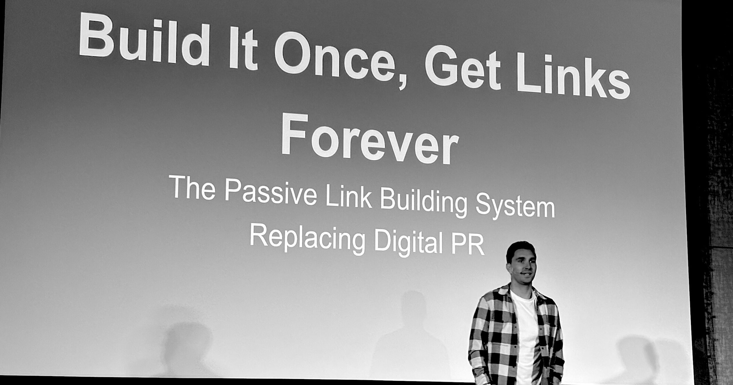 A speaker presents a passive link building strategy on stage with a bold SEO marketing slide promoting long-term backlink generation and digital PR alternatives.